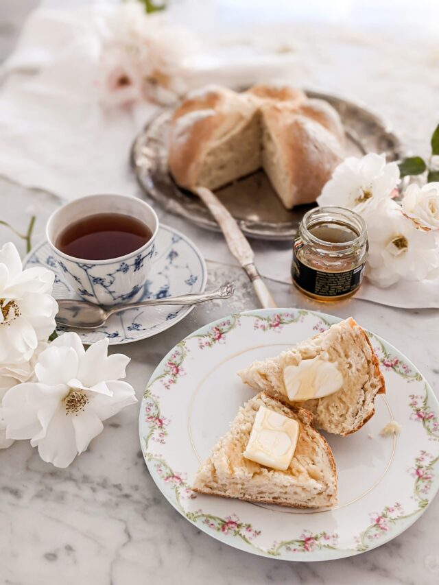 In the foreground is a floral plate with fresh bread and a teacup full of tea. Blurred in the background is the rest of the loaf of bread.