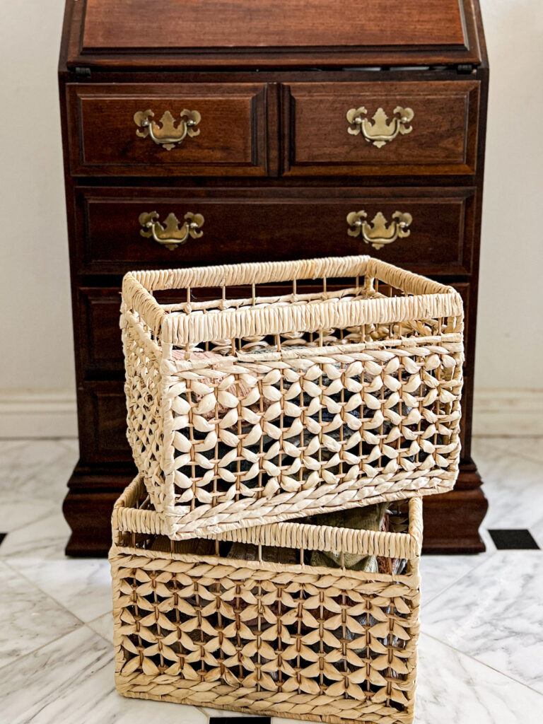 Two banana leaf baskets from Ikea are stacked in front of a chest of drawers with brass Chippendale-style handles.