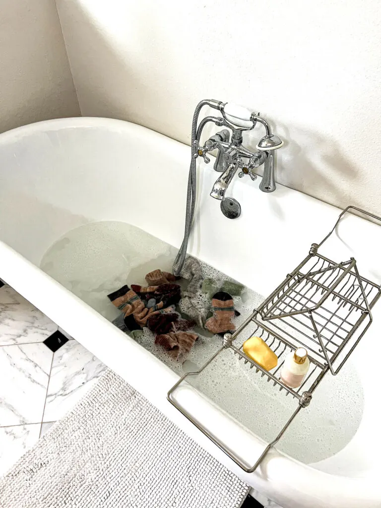 A white clawfoot tub with chrome tap and metal tray is partially filled with bubbly water. In the middle of the tub is a pile of wet socks jumbled together, soaking in the water.