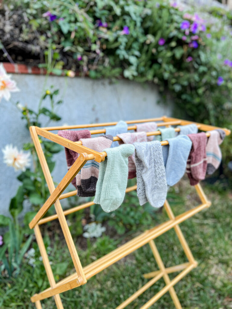 A lower angle view of the left side of a bamboo drying rack full of pastel socks in a garden. Blurred in the background are lots of flowers and creeping vines.