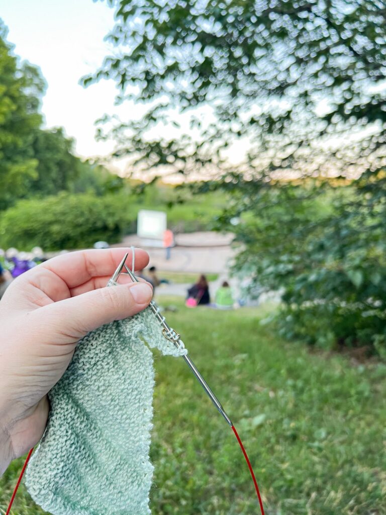 A white woman's hand holds up a light green shawl in progress on the knitting needles. Blurred in the background is lots of greenery and a speaker giving a presentation to a small crowd.