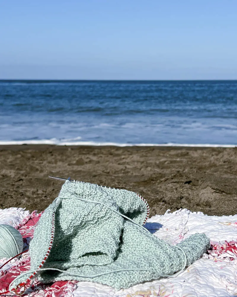 A light green knit shawl in progress sits on a floral blanket on the beach. Blurred in the background are soft ocean waves.