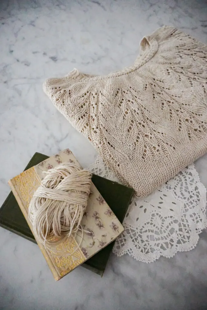 A knit linen t-shirt with a round lacy yoke is folded neatly with the sleeves and hem tucked under. It sits on a white marble countertop. In the foreground are a couple antique books with a ball of leftover linen yarn sitting on top.