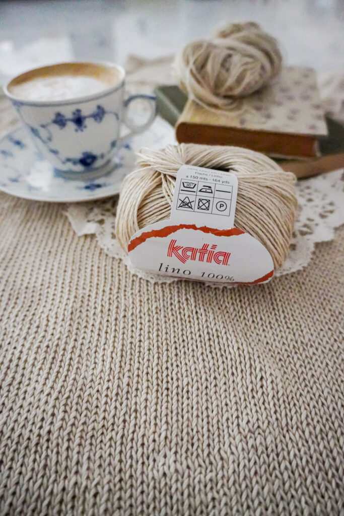 A close-up on the fabric of a knit linen tshirt. Sitting on top of it and still in focus is another ball of the same yarn with the label still attached (Katia Lino 100%). Slightly blurred in the background are a blue and white teacup full of coffee, some antique books, and more yarn leftovers.