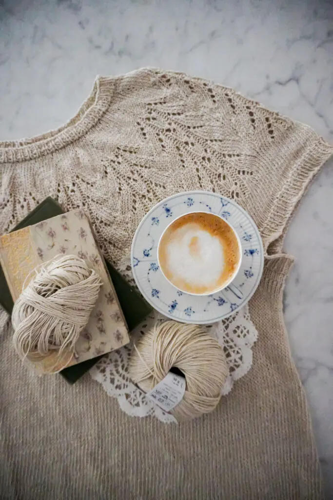 A top-down photo of a knit linen t-shirt with a lacy round yoke. Sitting on top of the t-shirt are a blue and white cup filled with a foamy latte, a couple antique books, and more balls of linen yarn.
