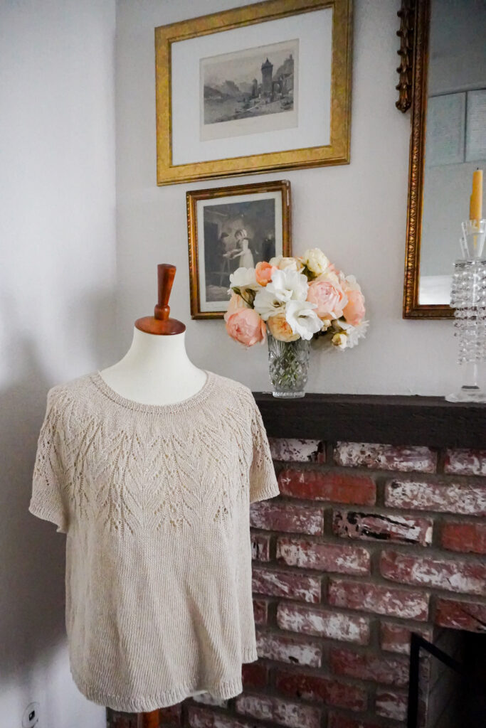 A tan linen shirt knit with a round lacy yoke is displayed on a white dressmaker's form with a wooden top. It stands in front of a brick fireplace. there is a vase of roses on the mantel and there are antique prints on the wall.