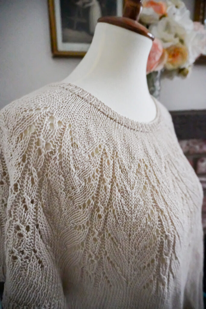 A close-up on the lacy yoke of a knit linen t-shirt. The shirt is displayed on a white dressmaker's form with a wooden top.