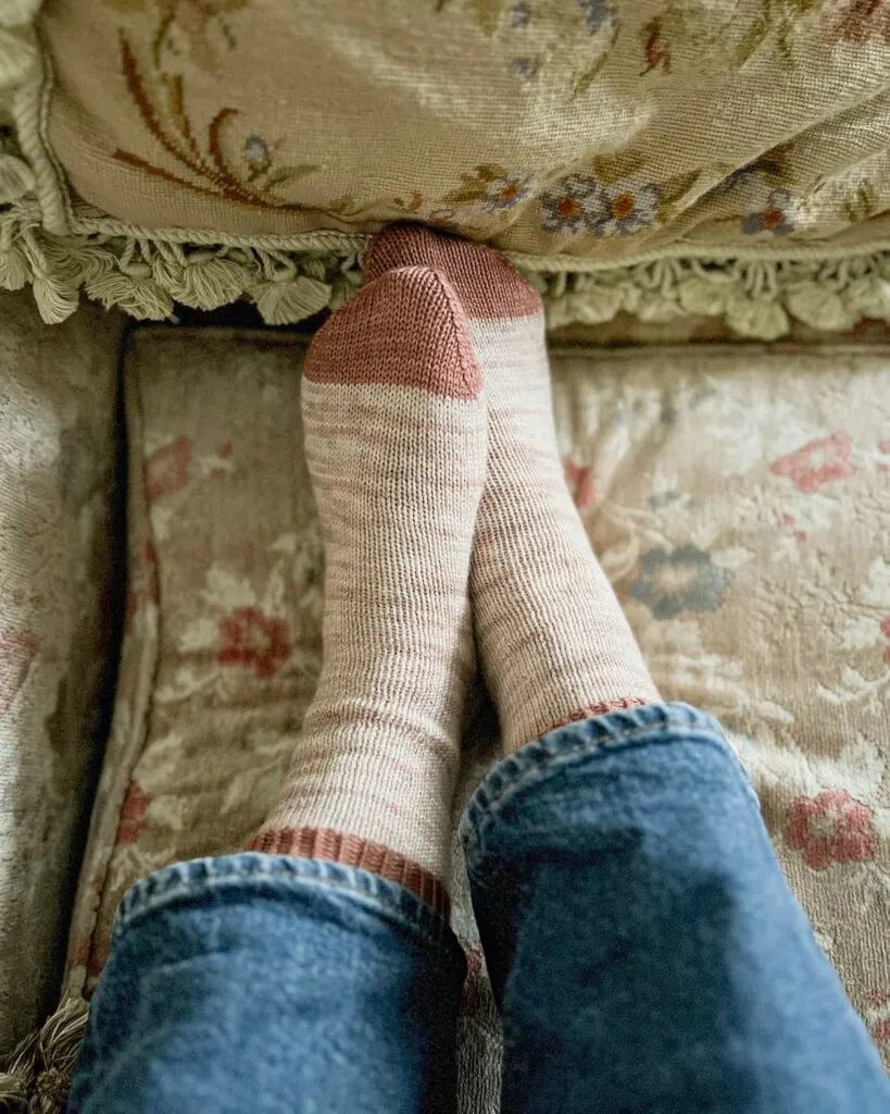 A pair of feet wearing softly variegated, pink, handknit socks with contrasting cuffs and toes are lightly crossed one over the other on a tan couch cushion with pink and blue flowers on it.
