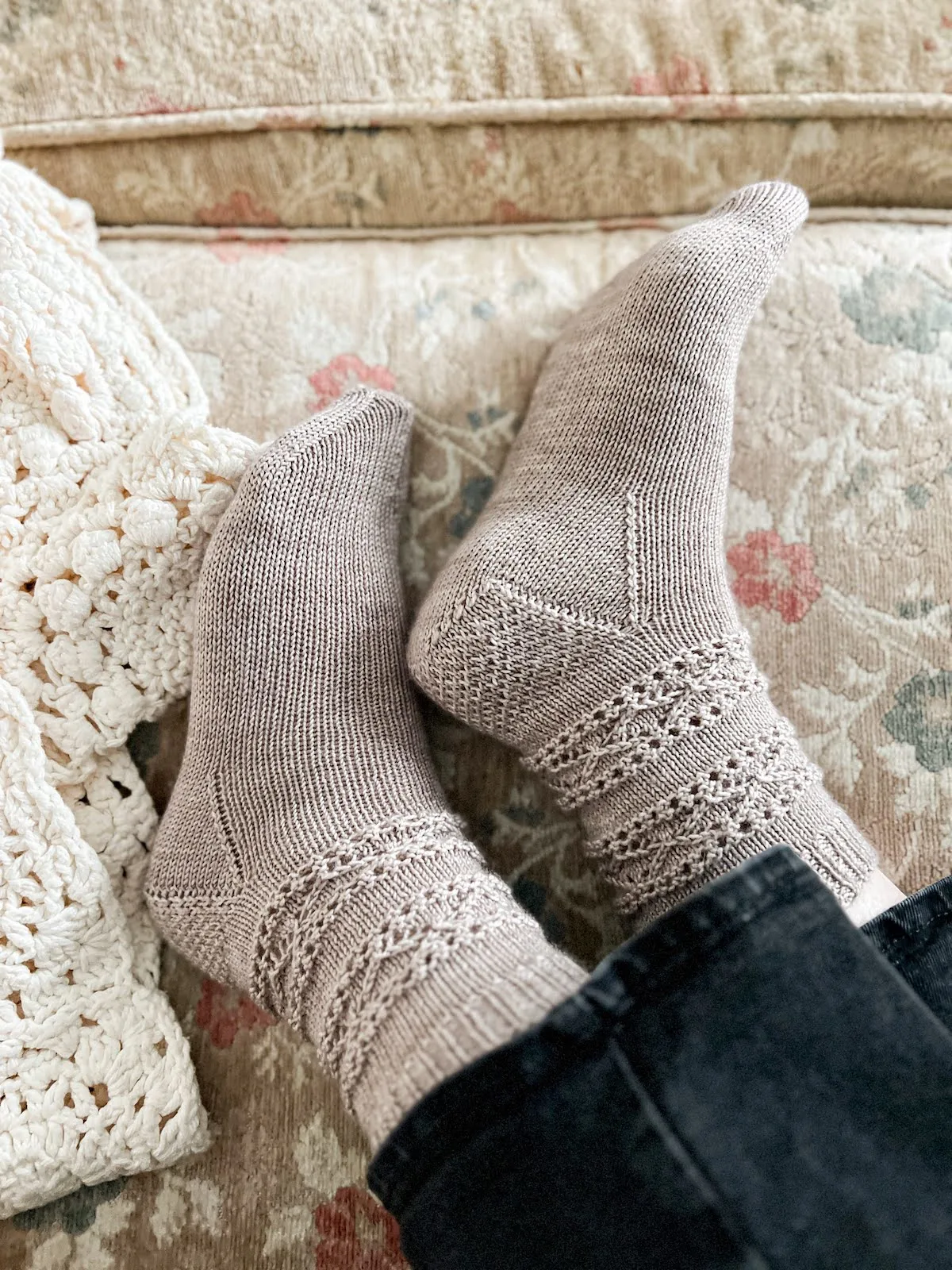A pair of feet wearing pearly gray handknit socks with a richly textured leg and eye of partridge heel are settled on their side against a tan couch cushion with pink and blue flowers on it.