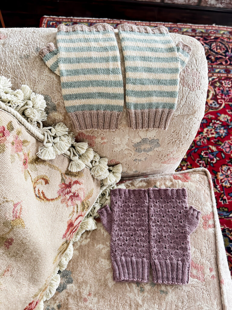 Two pairs of handknit mitts sit on the arm and cushion of a sofa with faded floral upholstery. The top pair is white and blue striped with contrasting gray trim on the cuffs and fingertips. The bottom pair is a textured purple pattern.