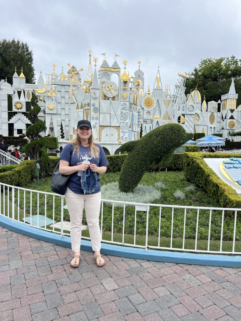 A blonde, white woman in her late 30s stands in front of the Small World ride at Disneyland. She’s wearing cream jeans, a Cinderella tshirt, and a black baseball cap, and she’s grinning at the camera. In her hands is a blue knit tshirt in progress.