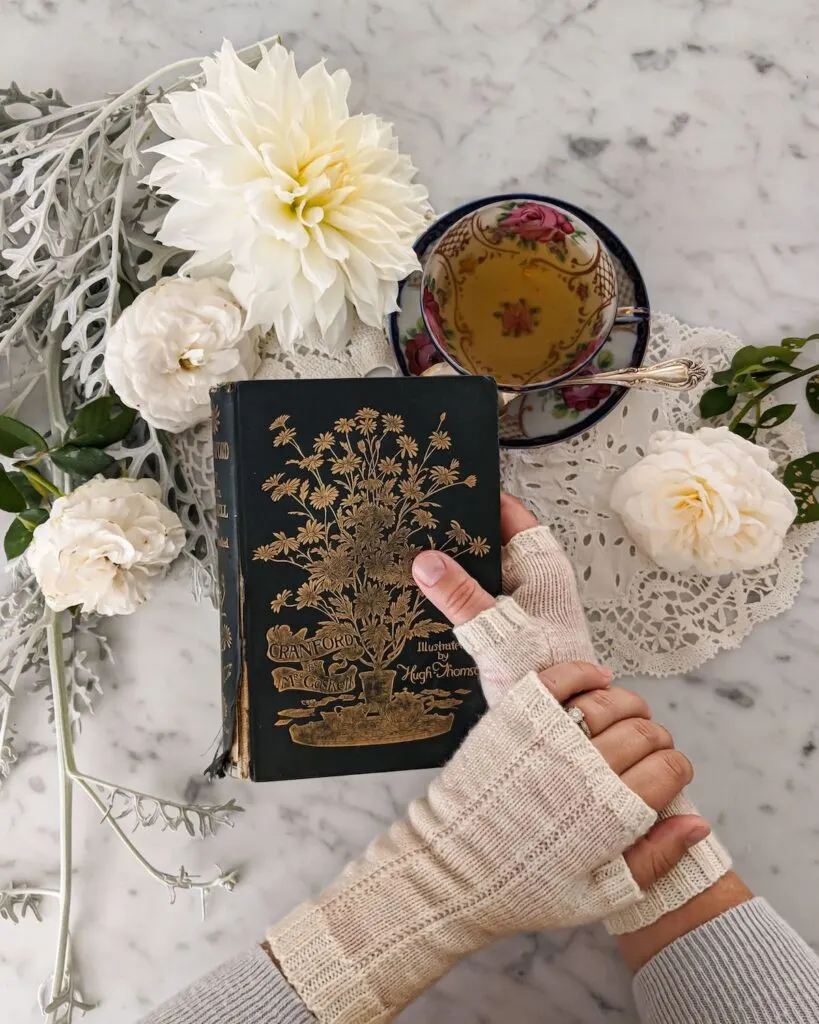 A woman's hands holds a dark green antique book with a gilded cover. On her hands are a pair of pale pink fingerless mitts. In the background is a teacup full of tea and some flowers.