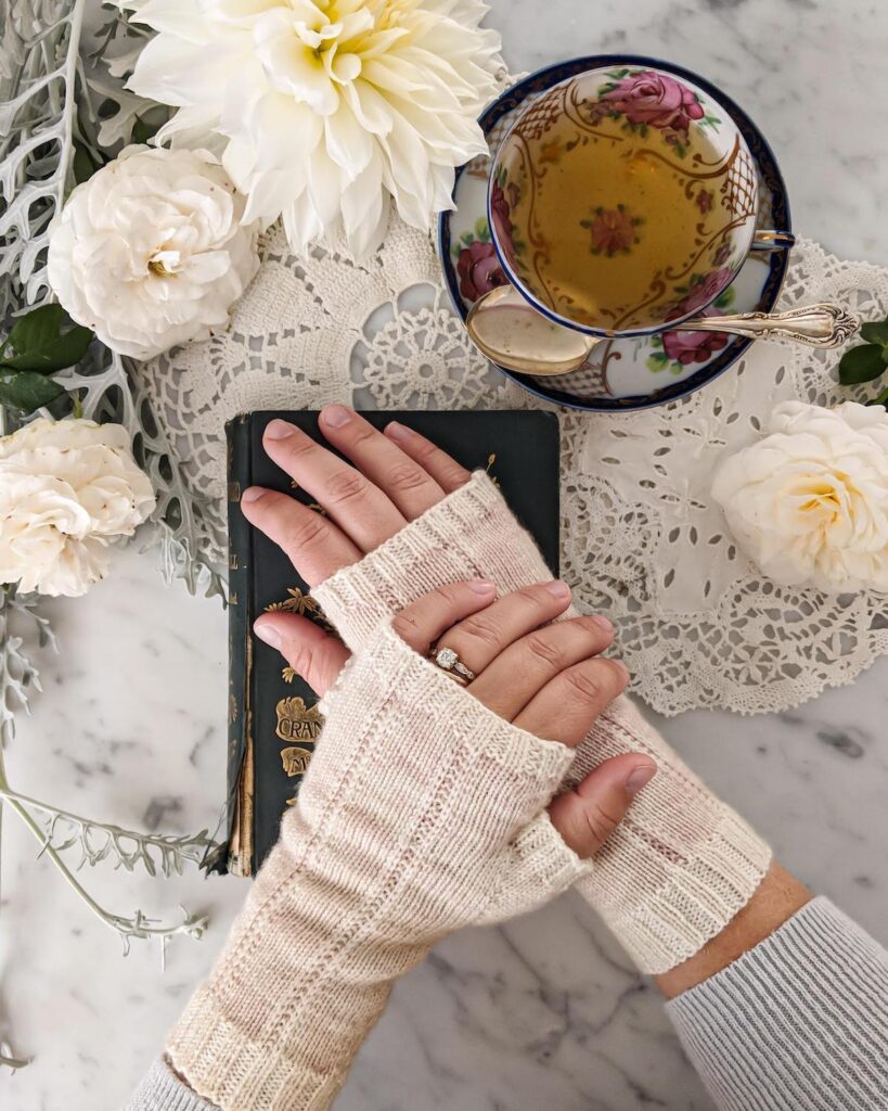 A woman's hands are crossed on top of an antique book, left over right, while wearing a pair of knit fingerless gloves. The mitts are pale pink with contrasting white trim.