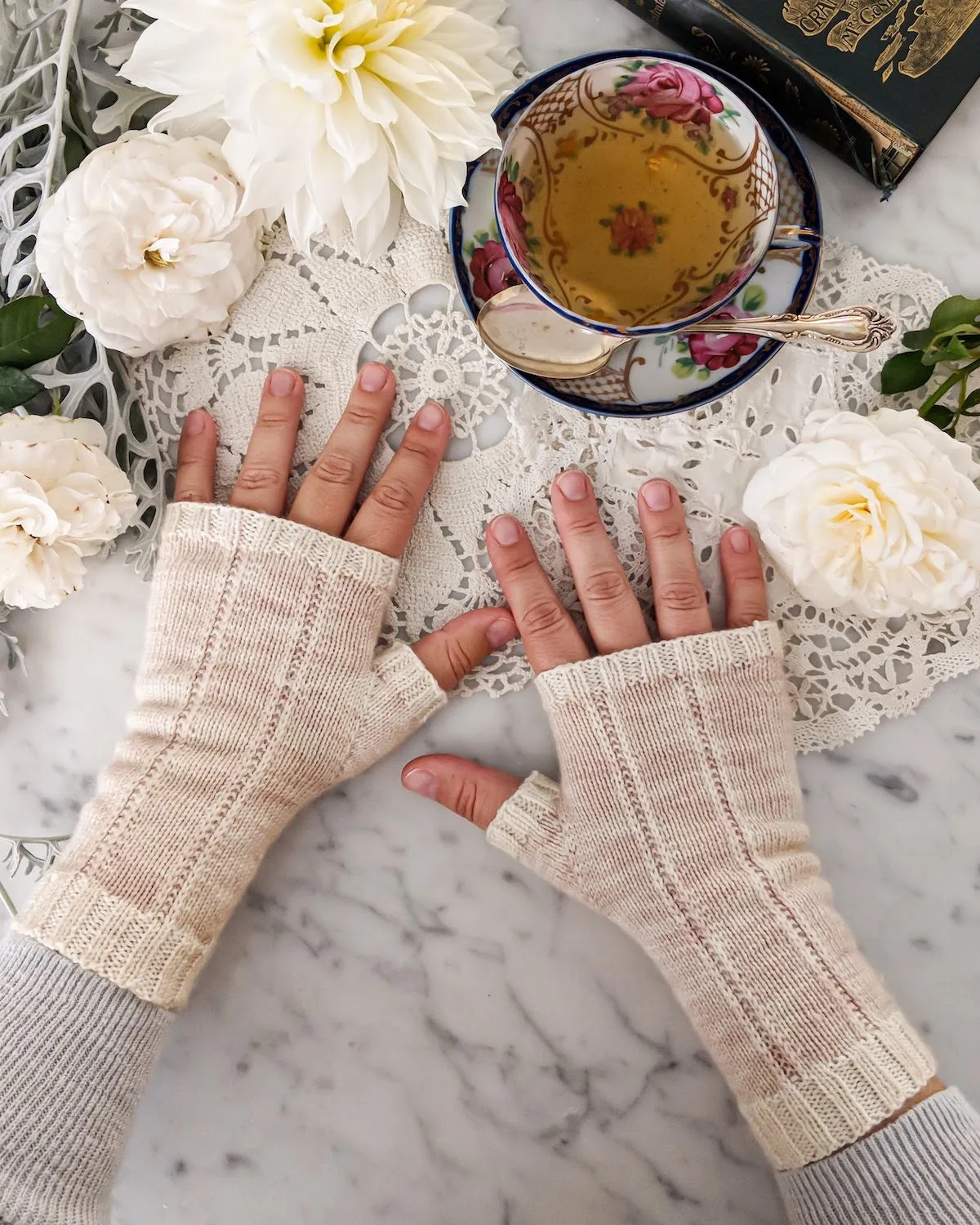 A woman's hands are splayed out on a countertop wearing pale pink fingerless mitts with cream contrasting trim. There are roses, dahlias, and a teacup full of tea, too.