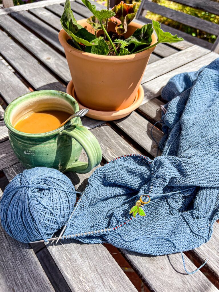A handknit sweater in progress on a wood slatted table in the sunshine. Next to it are a mug full of coffee and a pot of geraniums.