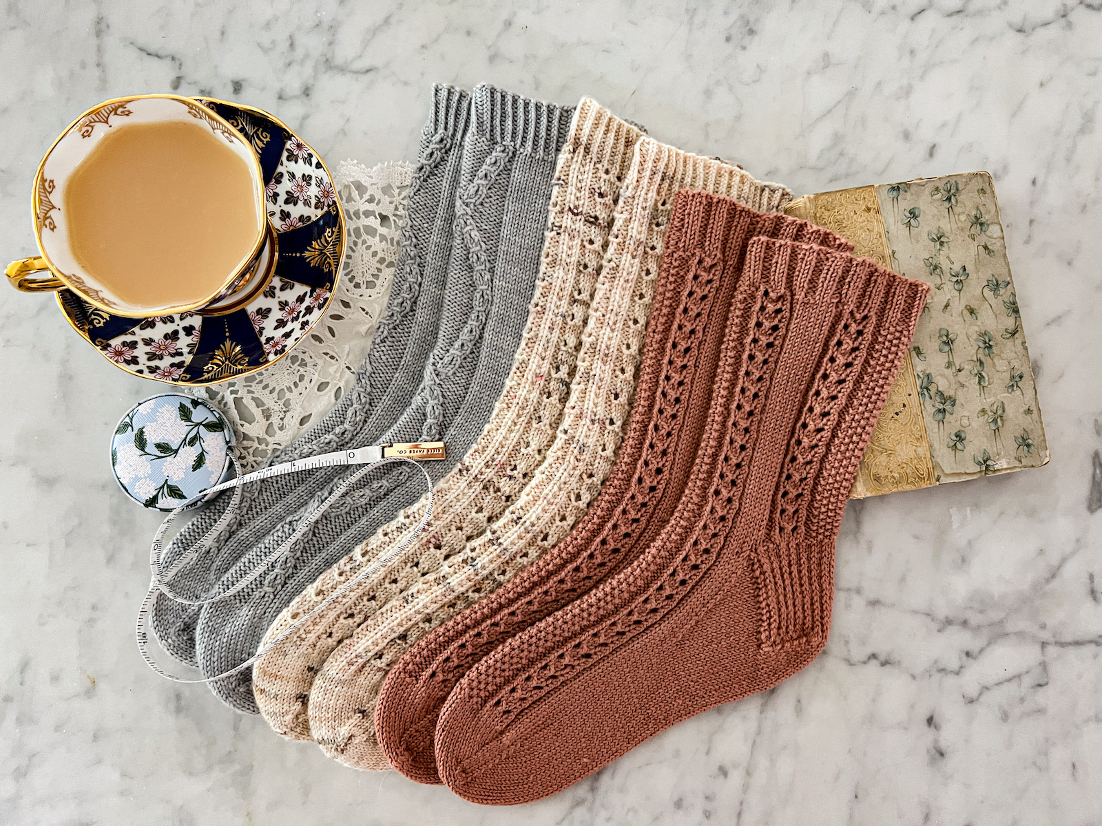 Three pairs of pastel handknit socks are laid out on a white marble countertop next to a teacup full of milky tea and an antique book. A little tape measure is spooled out across their toes.