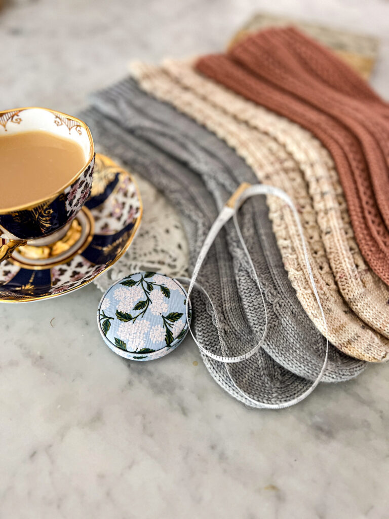 The foreground focuses on a little tape measure spooled out across the toes of three pairs of handknit socks. The rest of the socks blur away into the background. To the left is a teacup full of milky tea.