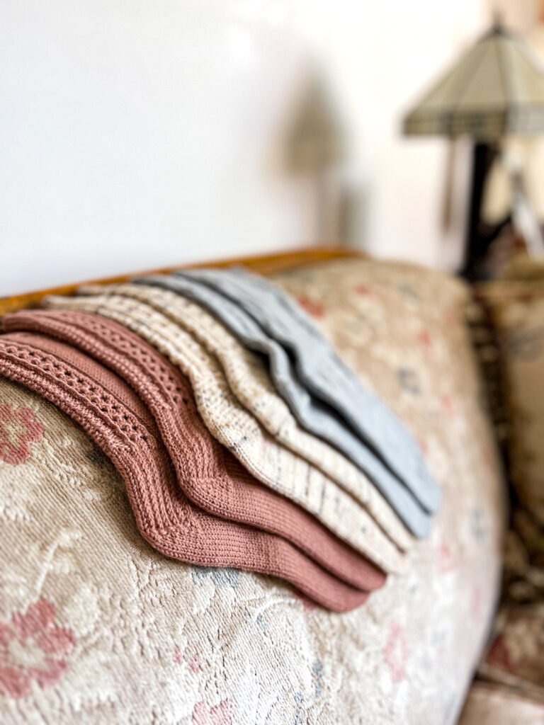 Three pairs of pastel handknit socks are laid across the back of an antique sofa with faded floral upholstery. The socks and the upholstery are color coordinated.