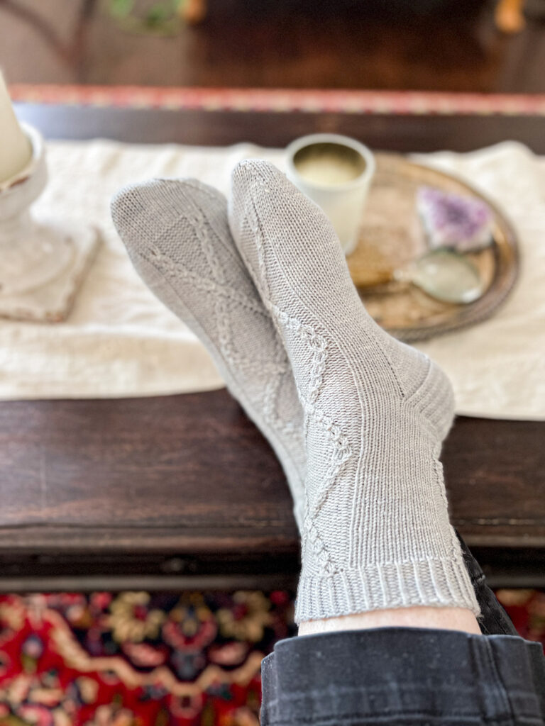 A pair of feet wearing light blue knit socks rest on a dark wood coffee table. The feet are crossed at the ankles.
