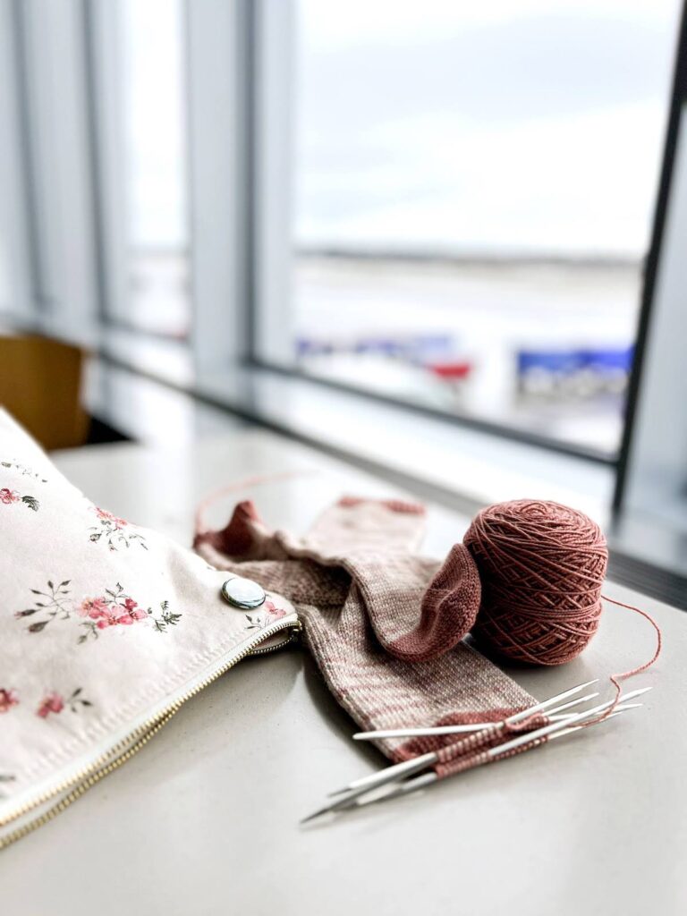 A softly variegated pink sock in progress sits on a table in front of a window at the airport in Portland, Oregon. I knit these socks while waiting for my flight home from Rose City Yarn Crawl 2023.