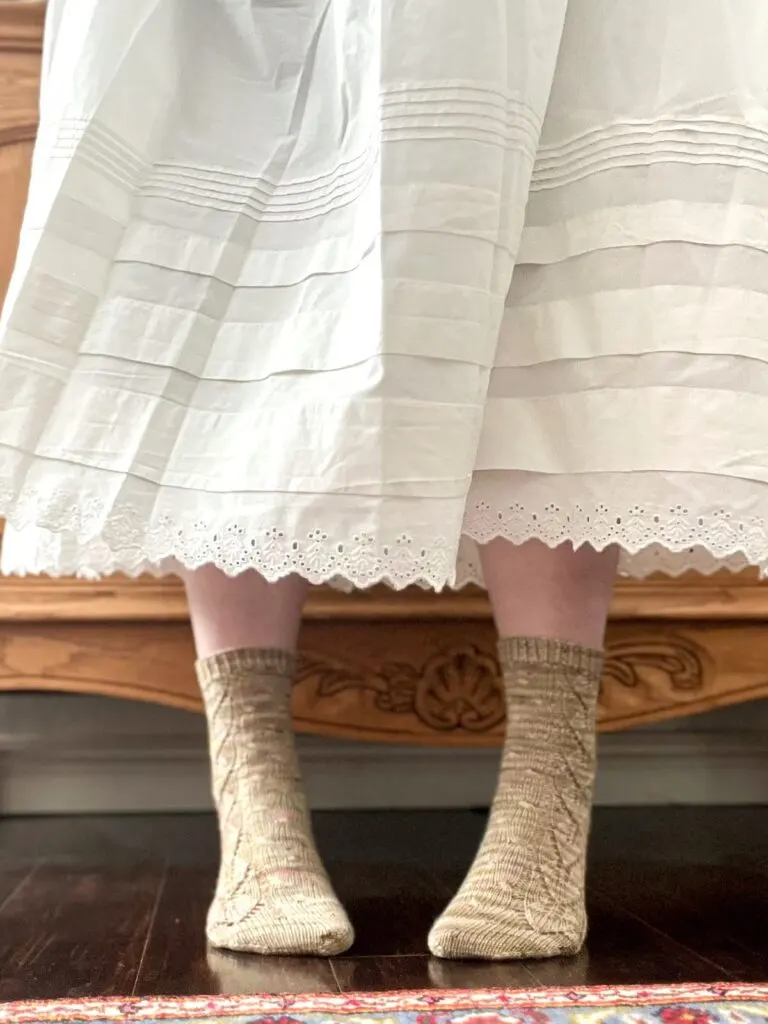 A pair of feet in handknit green speckled socks peep out beneath a white skirt with eyelet trim. The wearer is in the middle of a little twirl.