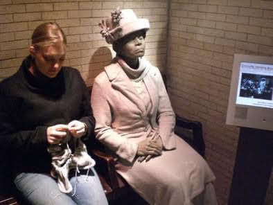 A white woman in her mid-20s sits next to a statute of another woman in an exhibit at the Smithsonian. The statue is wearing mid-20th century clothing and a cheery hat. The human woman is knitting.
