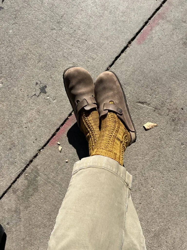 A pair of feet are crossed at the ankles on a sidewalk. They're wearing caramel-colored handknit socks and brown leather Birkenstock clogs.