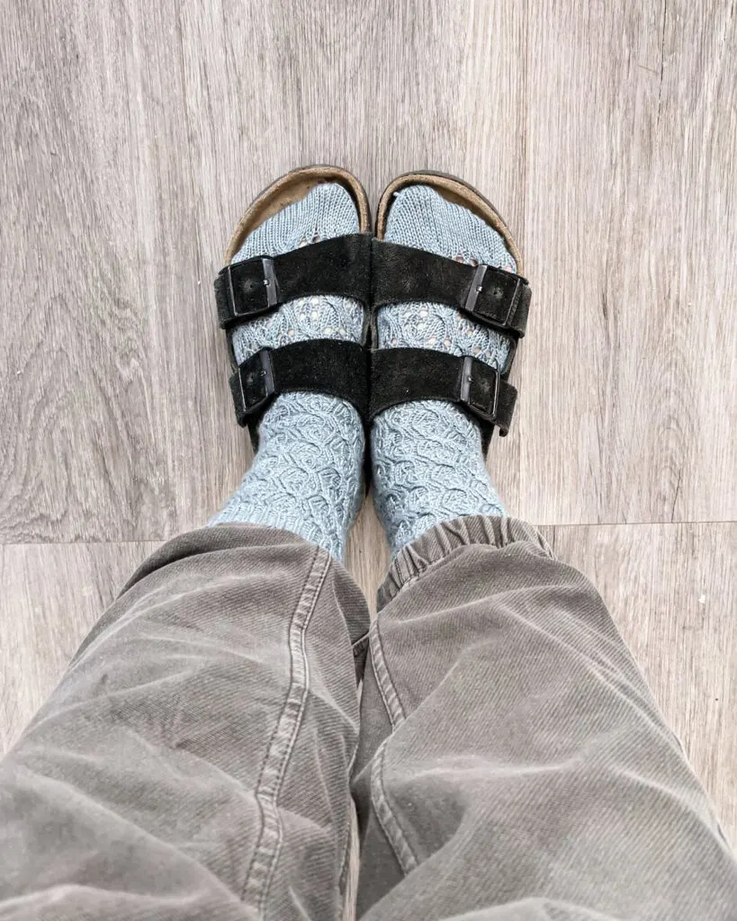 A pair of feet wearing light blue handknit socks and black Birkenstock sandals against a gray wooden floor.