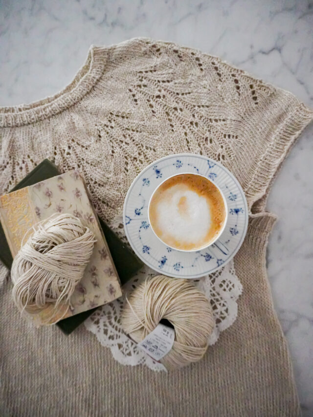 A top-down photo of a knit linen t-shirt with a lacy round yoke. Sitting on top of the t-shirt are a blue and white cup filled with a foamy latte, a couple antique books, and more balls of linen yarn.