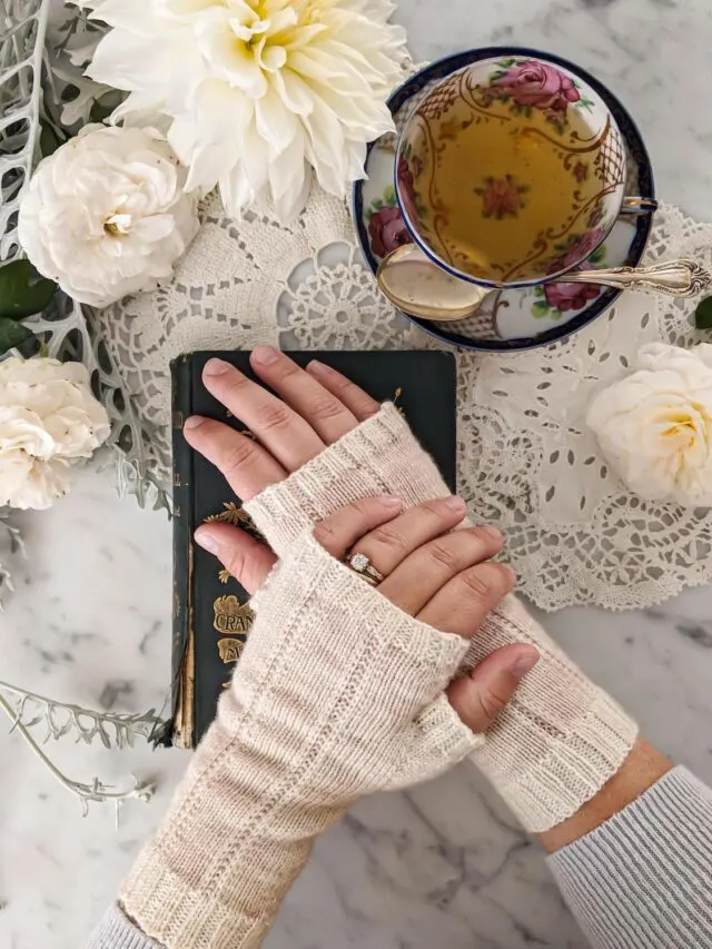 A woman's hands are crossed on top of an antique book, left over right, while wearing a pair of knit fingerless gloves. The mitts are pale pink with contrasting white trim.