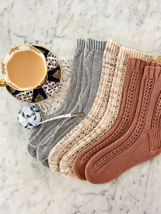 Three pairs of pastel handknit socks are laid out on a white marble countertop next to a teacup full of milky tea. A little tape measure is spooled out across their toes.