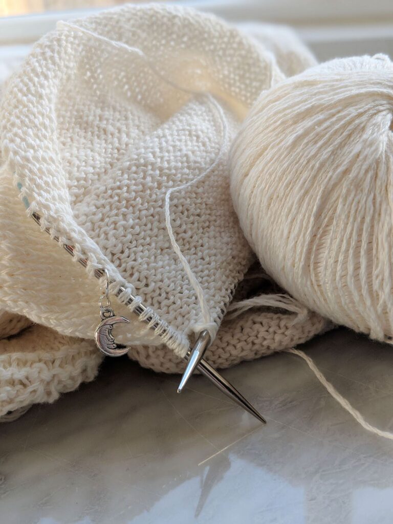 A white sweater sleeve in progress on a pair of metal circular knitting needles.