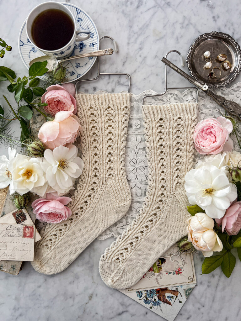 A pair of white handknit socks with a lacy texture is laid flat on a white marble countertop. The toes are both pointing to the left and the socks are displayed on wire sock blockers. They are surrounded by pink and white roses, a teacup full of Earl Grey tea, antique paper ephemera, a silver dish full of jewelry, and an antique silver curling iron.