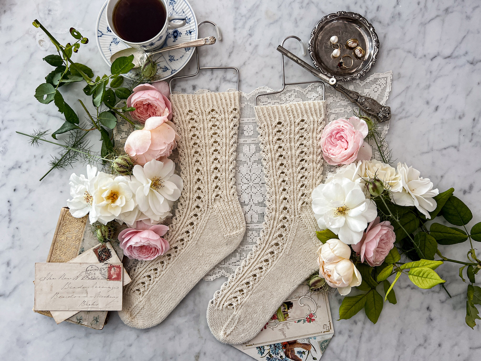 A pair of white handknit socks with a lacy texture is laid flat on a white marble countertop. The toes are both pointing to the left and the socks are displayed on wire sock blockers. They are surrounded by pink and white roses, a teacup full of Earl Grey tea, antique paper ephemera, a silver dish full of jewelry, and an antique silver curling iron.