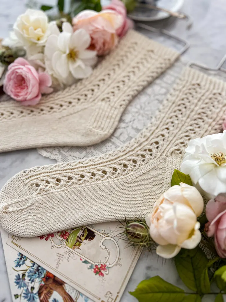 The foreground of the photo shows the foot of a pair of white handknit socks with a lacy texture. The leg and the other sock blur in the background. The socks are surrounded by pink and white roses and antique paper ephemera.