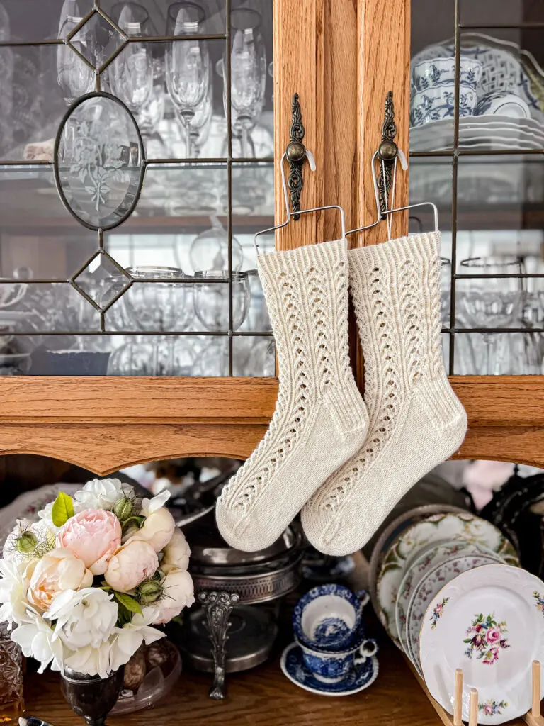 A pair of white, lacy, handknit socks hangs from the knobs of an oak china hutch. Below the socks are a vase full of roses and lots of antique dishes.