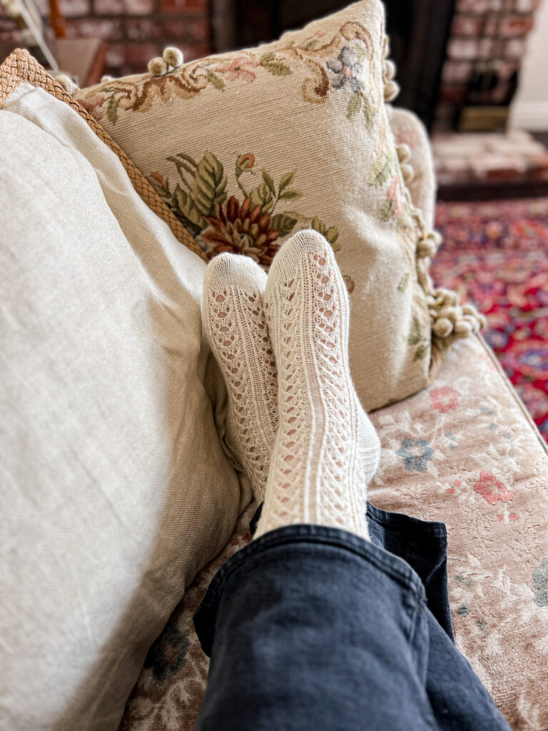 A woman's feet wearing a pair of white, lacy, handknit socks are crossed at the ankles on a soft pastel sofa.