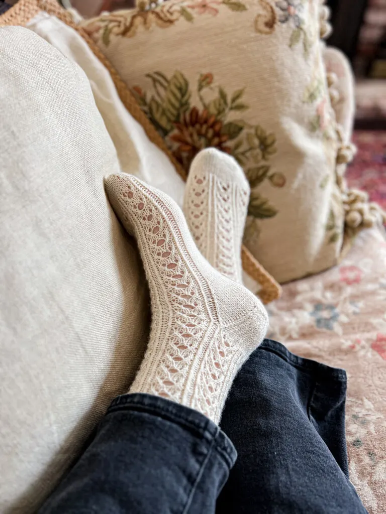 A woman's feet wearing a pair of white, lacy, handknit socks are stretched out one on top of the other. The left foot, on top, is rotated slightly to show the heel details of the sock.