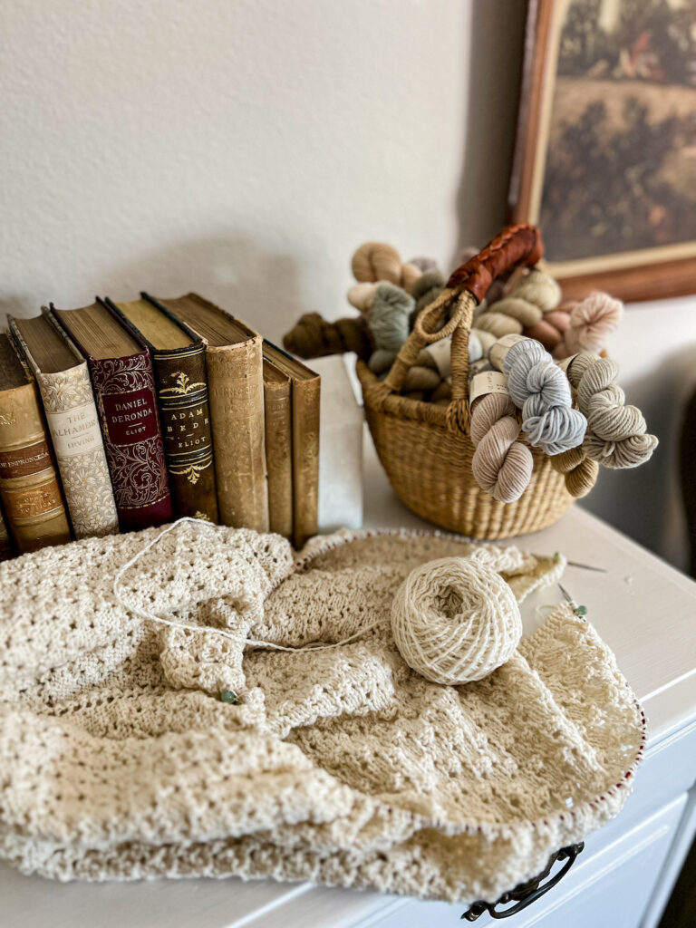 A white knit shawl in progress piled next to a small basket of mini skeins of yarn and several antique books.