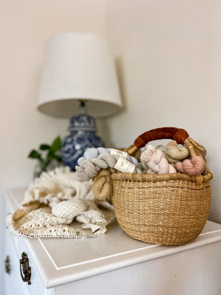 A grass basket full of mini skeins of yarn. The skeins are all pastel colors and spilll over the side like flowers in a bouquet. Blurred in the background are a white knit shawl on the needles and a blue and white lamp.