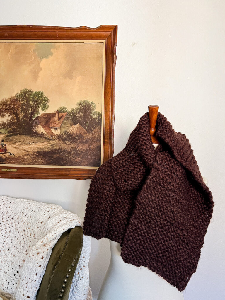 A dressmaker's form draped with a dark brown garter stitch scarf. In the background is a print of an old landscape painting in a wooden frame.