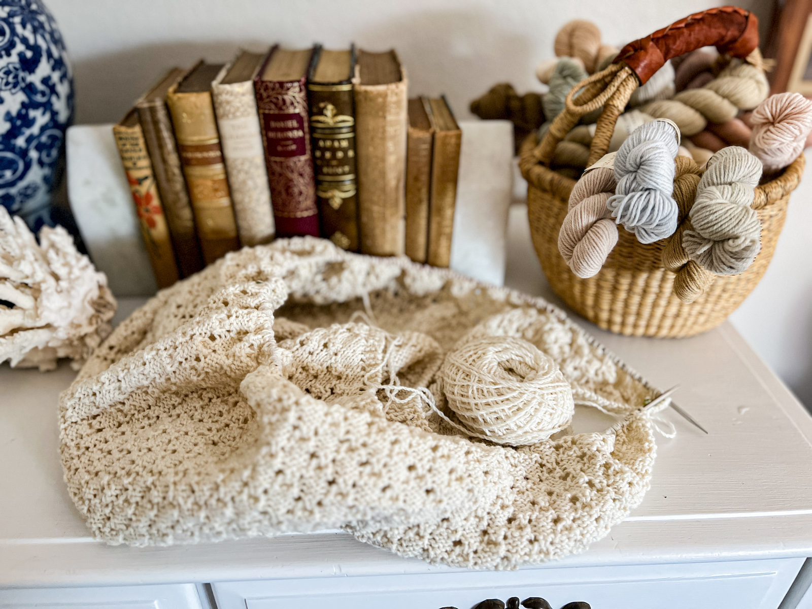 A white knit shawl in progress is piled on top of a dresser. Slightly blurred in the background are lots of antique books and a basket full of mini skeins of yarn.