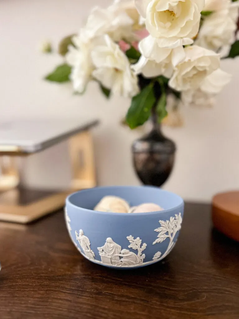 A small jasperware bowl sits on a dark wood surface. A couple small balls of yarn are barely visible over the rim of the bowl. Blurred in the background are a vase full of flowers and a laptop on a stand.