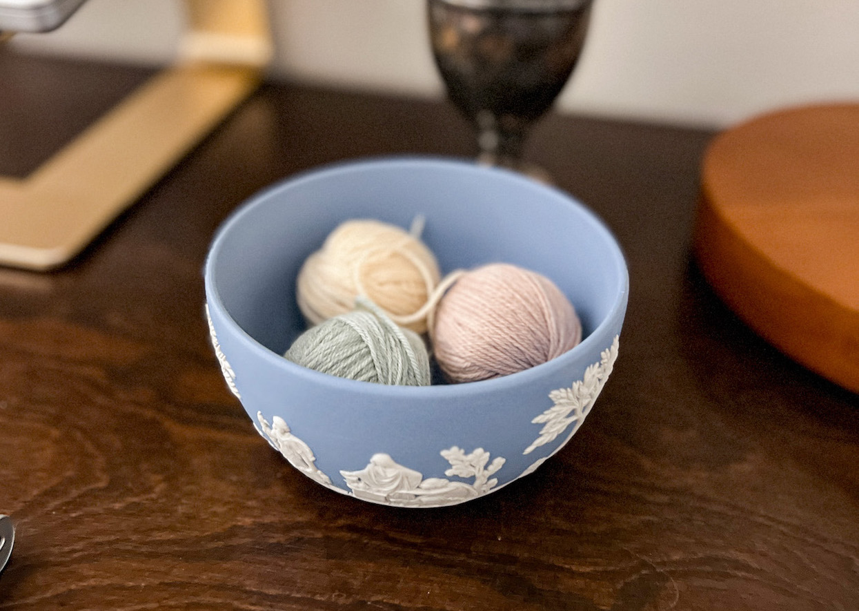 Three small balls of yarn sit inside a light blue jasperware bowl on a dark wood desktop.