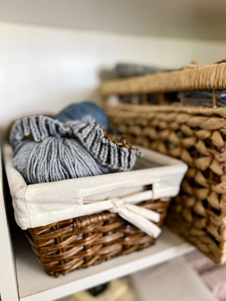 A wooden basket with a fabric liner sits on a white shelf. A knitting project in progress, with blue and brown brioche stitches, sits inside the basket.