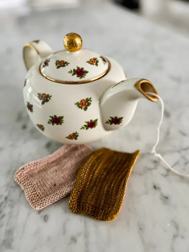 A floral teapot with a strand of pink yarn coming out of the spout sits on a white marble countertop next to pink and caramel-colored yarn swatches.