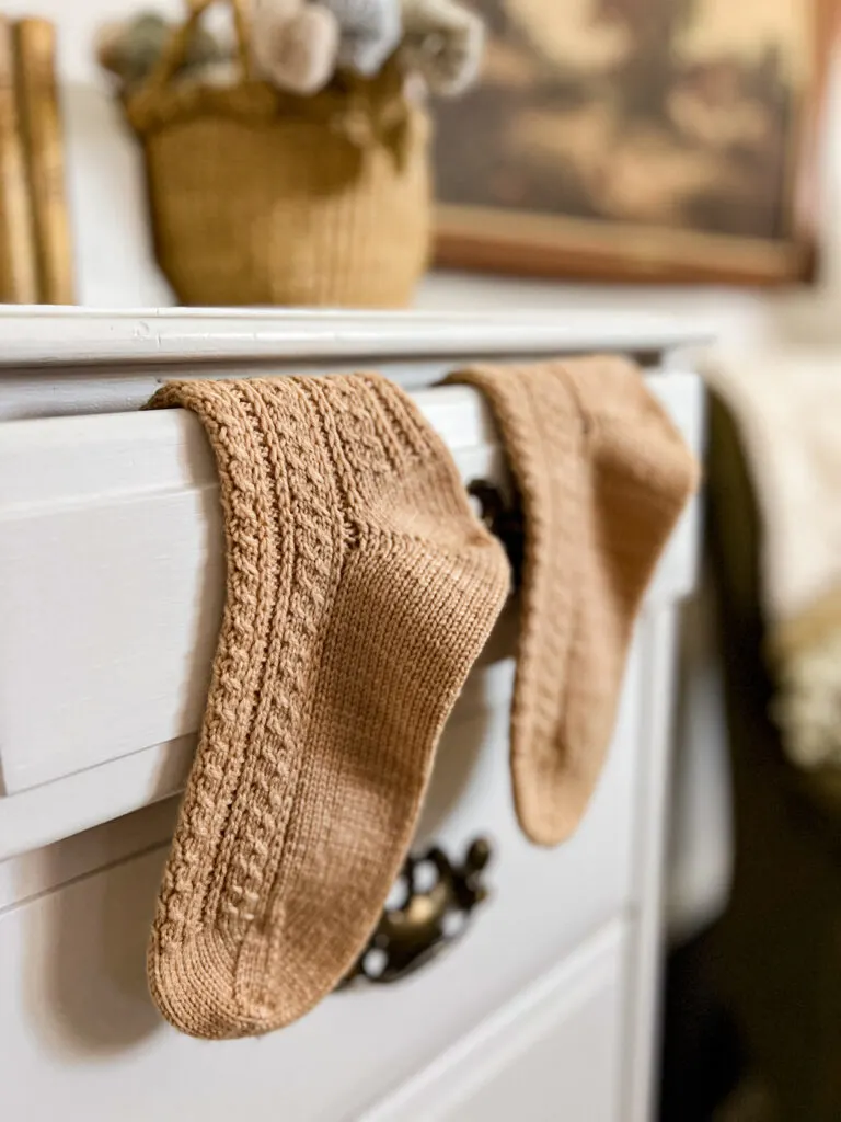 A pair of peach hand-knit socks with lots of tiny cable stitches peeks out of the top drawer of a white dresser.