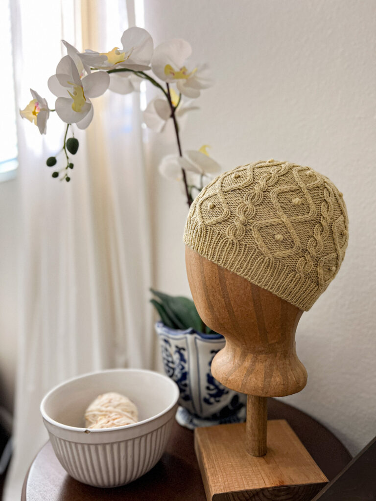 A light green knit hat is displayed on a brown head mannequin. The knit hat is full of cables and bobbles. It sits next to a planter with a fake orchid in it and a bowl full of yarn.