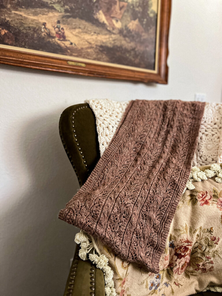 A brown knit wrap is draped across a green wingback chair and a needlepoint pillow. The wrap has lots of cables running up and down its entire length.