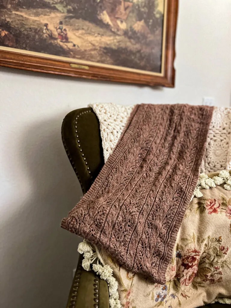 A brown knit wrap is draped across a green wingback chair and a needlepoint pillow. The wrap has lots of cables running up and down its entire length.